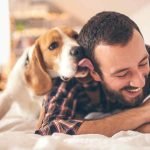 A dog licking an owner's ear who's smiling on a bed