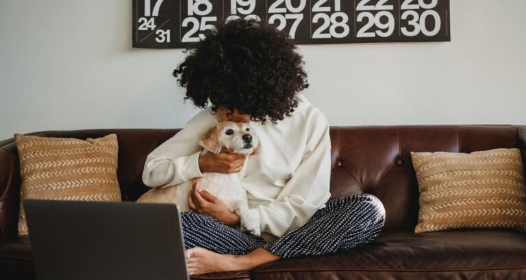A woman sitting on a couch is kissing a dog on the top of their head