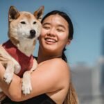 A woman is holding a dog wearing a harness outside