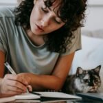 A woman is writing in a notebook while sitting on a couch with their cat